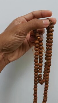 Vertical close-up shot of a person's hand holding and counting traditional brown wooden Muslim prayer beads (tasbih) against a plain white background.