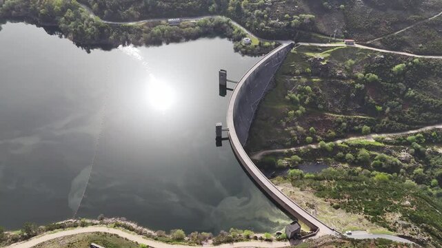 An aerial drone shot panning right over the Barragem de Vilarinho das Furnas dam and reservoir in a lush, hilly landscape near Campo do Ger&ecirc;s, Portugal. The sun reflects on the tranquil water.