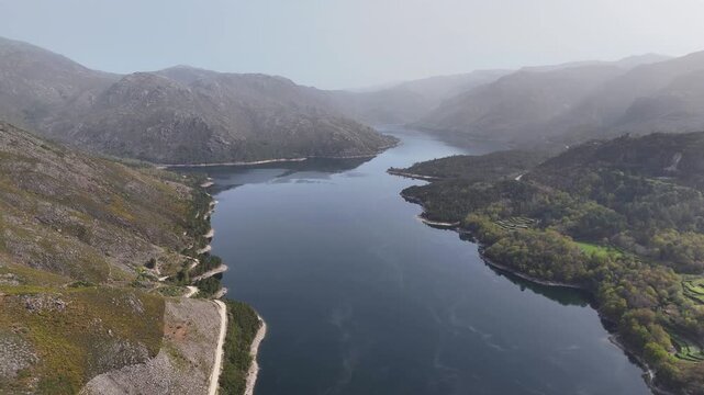 A drone pushes in over the scenic Picota mountain and a vast lake in the rugged landscape of Portugal. Lush green valleys and winding roads surround the tranquil waters under a sunny sky.