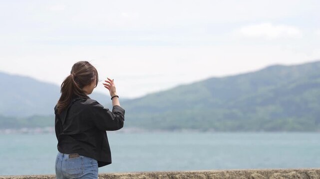 Woman stands by water, gazing at distant mountains. She holds her hand, as if touching the sky. Soft clouds drift above gentle hills. Her posture suggests quiet reflection
