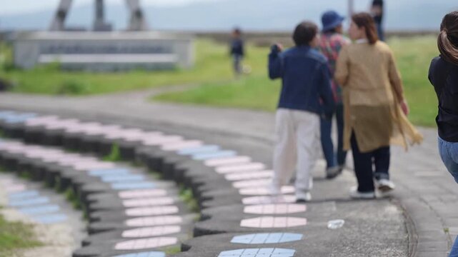 Woman walks with cane on paved path. Group of people strolls behind her. Blurry background shows open green space. Her expression suggests calm focus