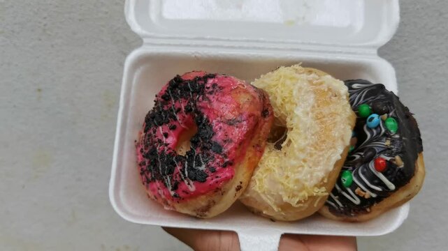 Top-down vertical shot of a hand holding a white styrofoam container filled with three different decorated donuts featuring pink glaze with chocolate crumbs, white cheese shavings, and chocolate glaze