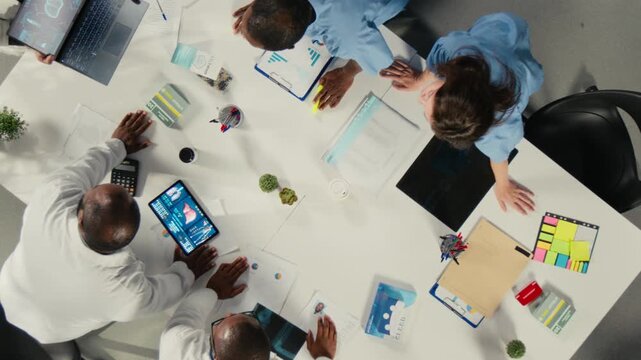 Doctors and nurses take part in a clinical discussion from an overhead angle inside a clinic meeting space, combining diagnostic data and treatment notes while they coordinate healthcare decisions.