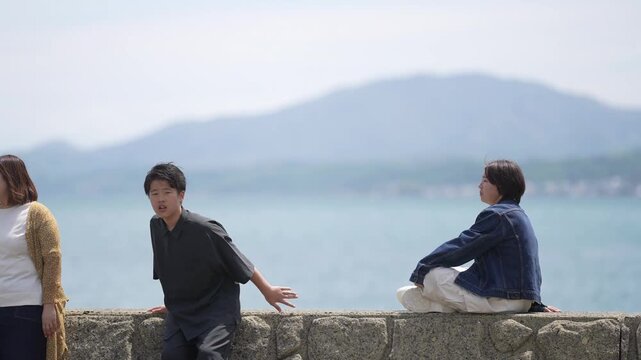 Two boys sit by the sea, one leaning, one gazing. They face each other, but minds drift apart. Sunlight warms the stones, sea hums in silence. Emotions linger, unspoken, gentle, quiet