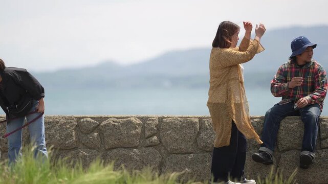 Three people relax near a seaside stone wall. One person leans forward with a fishing rod. Another sits on the wall, watching the water. Third gestures upward, perhaps in thought or joy