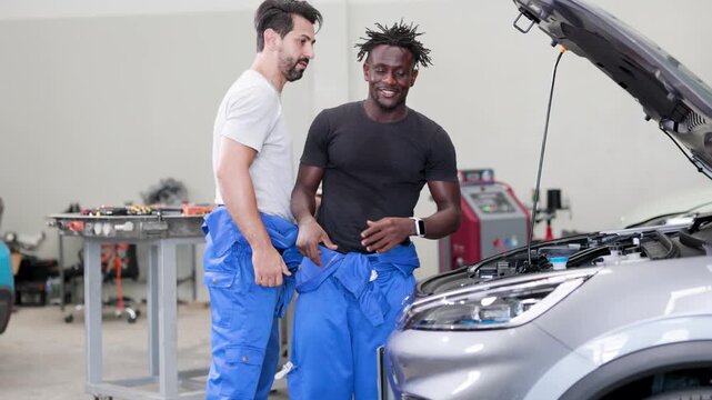 Two auto mechanics in blue overalls share a friendly handshake beside an open car hood in a workshop, celebrating teamwork and successful vehicle repair.