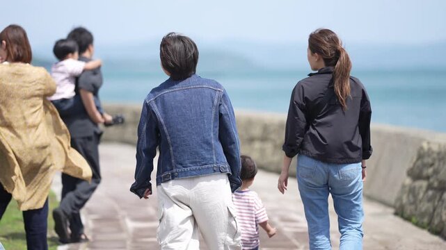 Family strolls seaside promenade. Two adults walk with children. Ocean waves roll gently. Children run ahead with joy. Sunlight reflects off water. Perfect for family vacation scenes