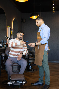 African American men exchanging smartphone and card reader in barbershop, barber wearing apron
