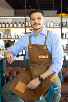 African American man sitting on high stool at cafe counter wearing brown apron, shelving jars