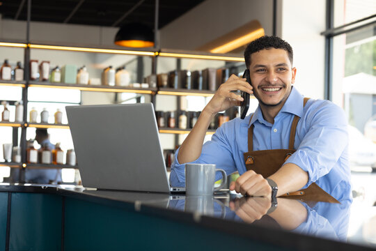 Man sitting at cafe counter wearing brown apron holding phone, smiling toward laptop and mug