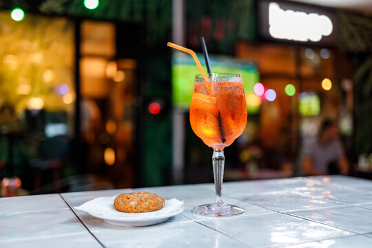 Orange cocktail beside round cookie on tiled bar table with colorful evening bokeh and softly blurred guests creating warm social nightlife atmosphere, with cocktail, cookie, bar, table detail