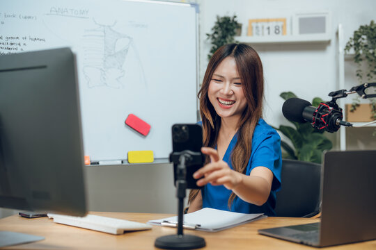 Asian female medical student broadcasting health podcast with microphone and smartphone. Young nurse in blue scrubs creating e-learning content and virtual anatomy class in modern home studio setup.