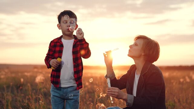Focused son with mother blows soap bubbles standing on meadow with growing plants at back sunset. Mother with son walking and blowing soap bubbles. Son and mother blow bubbles spending time together