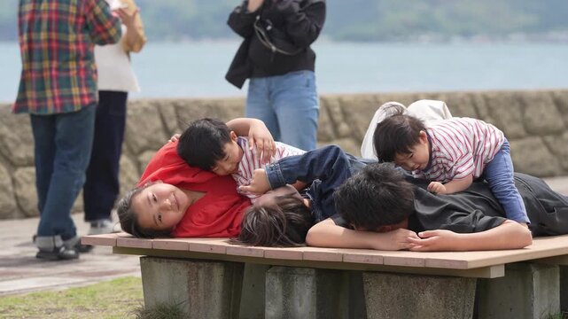 Children rest on a park bench near water. They lie close, heads together, smiling. Background figures observe without interrupting. Scene captures peaceful, joyful sibling bonding