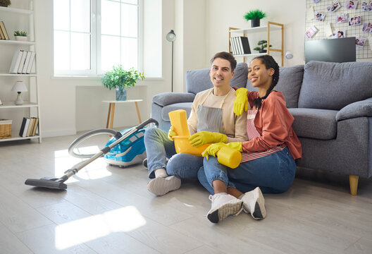 Couple cleaning with vacuum beside sofa, resting. Wearing gloves and holding a sponge, they pause on the floor after housework, smiling in a bright home interior. Concept of household teamwork.
