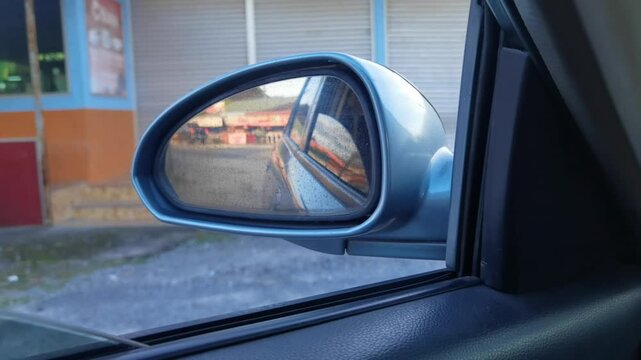 View of a car's side-view mirror reflecting a quiet street and closed storefront shutters on a sunny day.