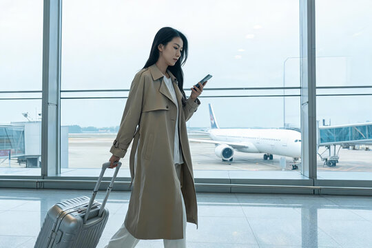 An asian woman walks in an airport terminal, pulling a rolling suitcase and using a mobile phone, with an airplane visible outside the window.
