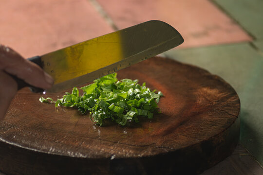 Close up of a hand holding a kitchen knife slicing fresh green culantro or herbs on a round wooden cutting board