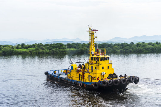 Harbor tugboat assisting a commercial vessel during port maneuvering with tropical mountains in the background at sunrise.