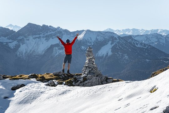 Mountaineer stretching his arms in the air, next to a cairn, in front of snow-covered mountains of the Rofan, hiking trail to Guffert with first snow, in autumn, Brandenberg Alps, Tyrol, Austria