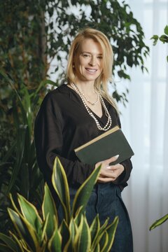 A middle-aged woman stands amidst vibrant indoor plants, holding a book close to her chest. She wears a black outfit and pearl necklace, showcasing a modern, elegant style