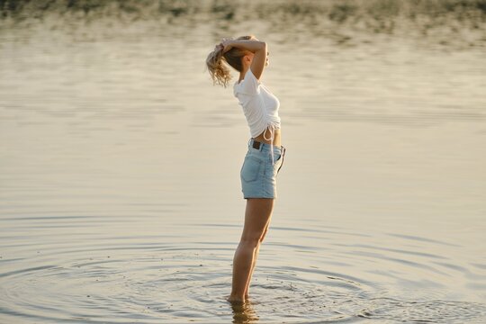 A woman stands in shallow water in profile, basking in the warm glow of sunset. She raises her hands and gathers her hair in a ponytail