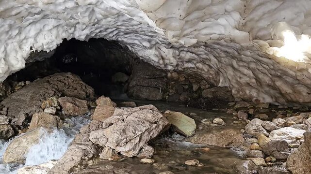 A rocky stream flows through a dark snow cave, with large boulders scattered along the water and layered snow forming an arched ceiling overhead.
📍 Hawraman Takht, Kurdistan, Iran 