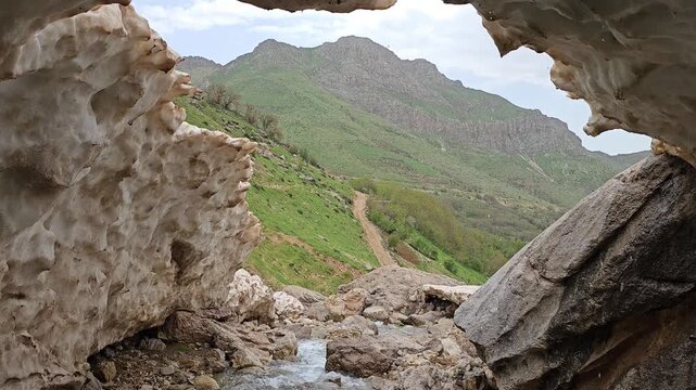 A rocky mountain stream flows out from beneath a snow cave, framed by boulders, with a dirt trail winding along green hillsides toward mountain peaks in the background.
📍 Hawraman Takht, Kurdistan