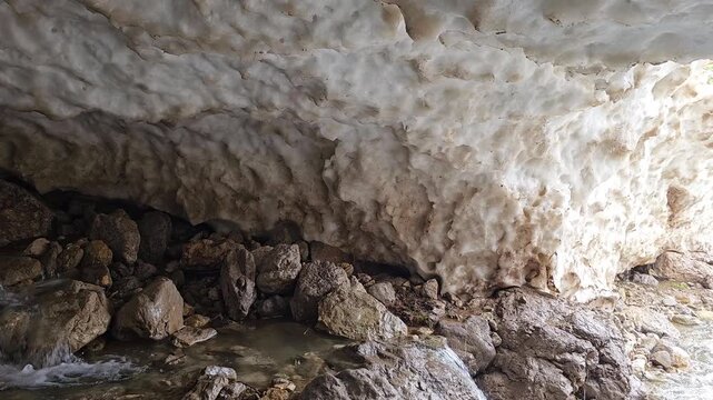 A rocky mountain stream flows out from beneath a snow cave, framed by large boulders, with a dirt trail winding along green hillsides toward mountain peaks in the background.
📍 Hawraman Takht