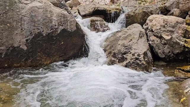 A rocky mountain waterfall cascades over moss-covered boulders near a snow cave, with patches of avalanche snow and rugged snow-capped peaks rising behind green hillsides.
📍 Hawraman Takht, Kurdistan