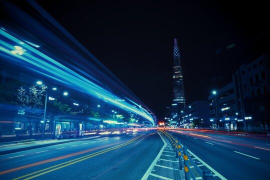 SEOUL, SOUTH KOREA - JUN 23, 2019: A futuristic blue-toned street view showing long light trails of city buses heading toward the distant Lotte World Tower under a night sky.