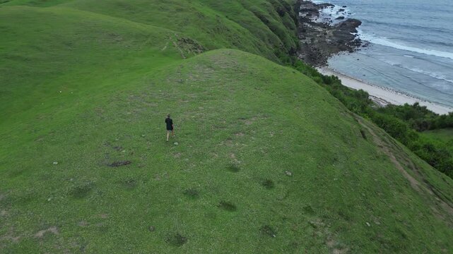 Woman Descending Coastal Cliff Path Toward Beach And Rocky Shoreline Narrow Trail, Surf Lines, Textured Grass Slopes, Overcast Light, Aerial Framing Conveys Journey And Scale, Bukit Merese, Lombok