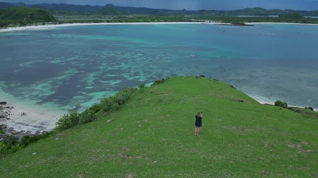 Woman Descending Coastal Cliff Path Toward Beach And Rocky Shoreline Narrow Trail, Surf Lines, Textured Grass Slopes, Overcast Light, Aerial Framing Conveys Journey And Scale, Bukit Merese, Lombok