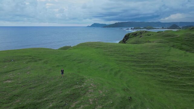 Woman Walking Across Rolling Coastal Hills With Dramatic Cloudscape Distant Shoreline Visible, Gentle Undulating Paths, Reflective Mood, Aerial Sweep Showing Scale And Textured,Lombok island,Indonesia