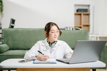 Fototapeta premium Asian woman manages business calls on her smartphone while working on a laptop with documents at home, blending corporate flair with cozy remote work.