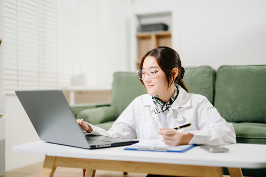 Asian businesswoman works from home, reviewing project financial documents with a laptop and coffee at a modern living room coffee table.