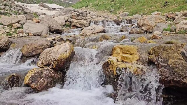 A rocky mountain stream cascades over moss-covered boulders, with patches of avalanche snow and rugged snow-capped peaks rising behind green hillsides.
📍 Hawraman Takht, Kurdistan, Iran 