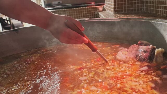 Chef mixes meat soup with vegetables in a large cauldron with a slotted spoon