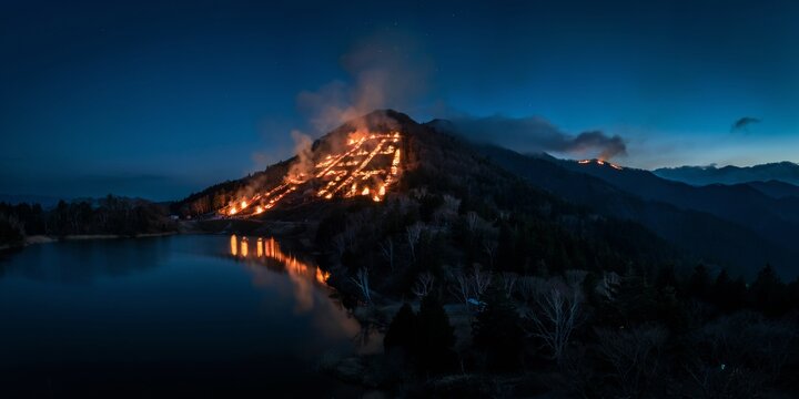 Large scale fire ritual on mountain slope forming japanese character dai during gozan no okuribi festival in kyoto japan reflected in calm lake water at night under dark blue twilight sky.