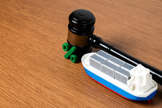 Miniature cargo ship, judge's gavel, and green percentage sign on a wooden desk. Represents import tariffs, maritime trade disputes, shipping taxes, customs duties.