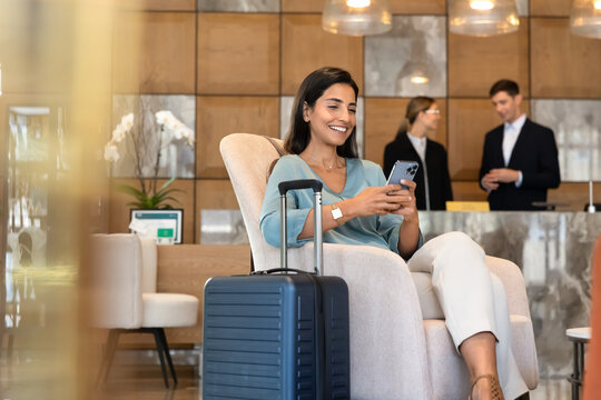 Female traveler guest sitting in hotel lobby, checking smartphone to read messages, confirm booking, browse travel information, using online customer services. Digital communication, hotel industry