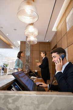 Receptionist assisting guest by phone call, working at front desk on computer, managing reservations, handling booking requests, providing support, answering inquiries, coordinate check-in, check-out