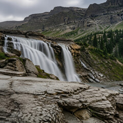 Obraz premium Majestic Cascading Waterfall Flowing Over Rugged Mountain Rock Formations in a Serene Alpine Wilderness Landscape