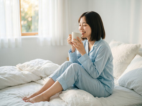 明るい寝室のベッドの上で湯気の立つカップを持ち微笑む女性