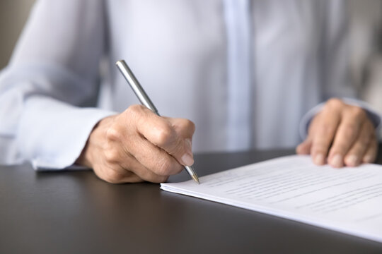 Signing agreement. Close up cropped shot female in white official shirt put signature by hand on paper document page. Making deal confirmation or sales purchase financial contract affirming concept