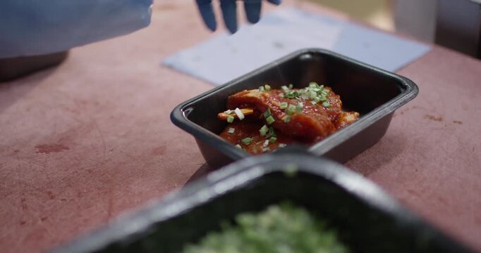 Chopped chives being sprinkled over marinated meat in a black tray with a bowl of fresh herbs in front