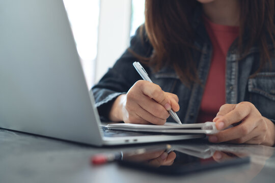 Woman hand with a pen writing in notebook with laptop on desk, doing online research, e-learning, online studying, business planning
