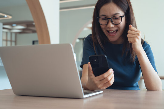 Excited cheerful young asian woman in casual dress sitting at table, using smartphone and laptop computer, raising hand up and celebrating success at home, close up