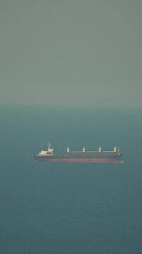 Industrial cargo vessel navigates across the deep horizon of a calm sea during a hazy afternoon. Fixed high-angle long shot.