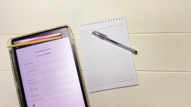 Top view of a male business owner holding a digital tablet and studying a business plan template for strategic business planning. Wearing eyeglasses with a notebook and pen beside the tablet